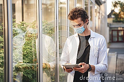 Young male scientist monitoring plant development with digital tablet in glasshouse during COVID-19 outbreak Stock Photo