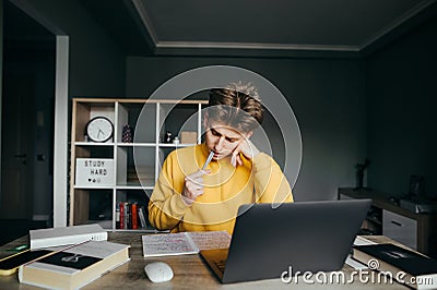 Young male student studying at home with books and laptop on bedroom background, looking at notebook with serous face and biting Stock Photo