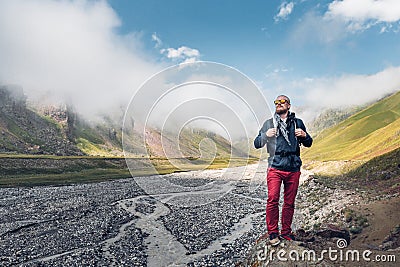 Young Male Traveler With Backpack Walks Along Valley Of A Mountain River Against The Background Of Mountains And Clouds Stock Photo