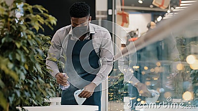 Young male waiter in apron wipes table sprays disinfectant cleaner prepares cafe restaurant for opening serious african Stock Photo