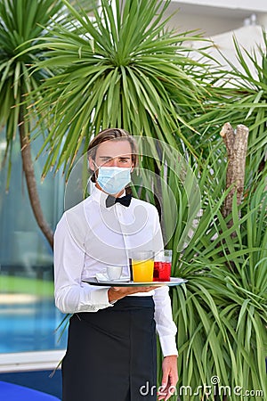 Young male waiter with a surgical mask holding a bar tray with assorted drinks outdoors on an out of focus background. Serving and Stock Photo