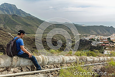 A young man with a backpack seats on the concrete aqueduct in the west side of Tenerife near the Adeje village. Hiking by the Stock Photo