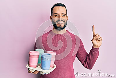 Young man with beard holding tray with take away coffee smiling with an idea or question pointing finger with happy face, number Stock Photo