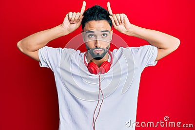 Young man with beard listening to music using headphones doing funny gesture with finger over head as bull horns Stock Photo
