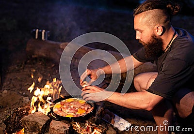 A young man with a beard prepares a dish of bacon with scrambled eggs on a fire. Cooking and outdoor recreation in summer Stock Photo