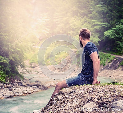 Young man with a beard and short hair, sitting on the bank of a mountain river in shorts and a blue shirt, with natural nature bac Stock Photo