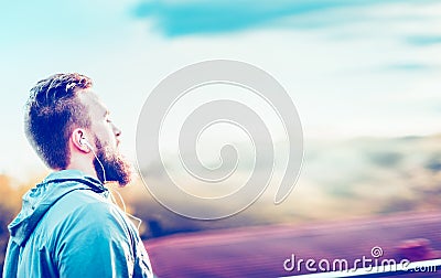 Young man with a beard and a short haircut, standing in profile against a blurred urban sunny landscape headphones in their e Stock Photo