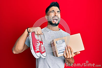Young man with beard taking casual red shoes from box angry and mad screaming frustrated and furious, shouting with anger looking Stock Photo
