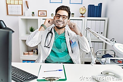 Young man with beard wearing doctor uniform and stethoscope at the clinic smiling pulling ears with fingers, funny gesture Stock Photo