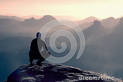 Young man in black sportswear is sitting on cliff's edge and looking to misty valley bellow Stock Photo