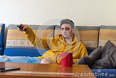 Young man, bored, watching the television while changing channels on it, laying in the sofa Stock Photo