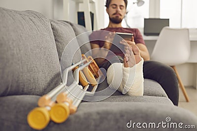 Young man with broken leg in cast sitting on couch with crutches and using tablet Stock Photo