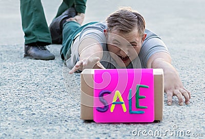 A young man buyer is lying on the sidewalk at the store and screaming with tension crawling to the box with the inscription sale. Stock Photo