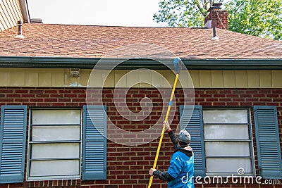 Young man cleaning the soffit of a one story house with a brush on a long pole. The house has blue shutters Editorial Stock Photo