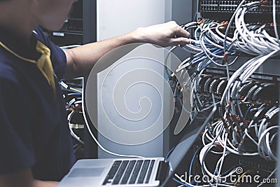 Young man connecting wires in server cabinet while working with laptop in data center Stock Photo