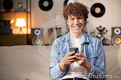 A young man in a denim shirt sits on the living room couch and smiles at the camera. The student boy holds a smartphone Stock Photo