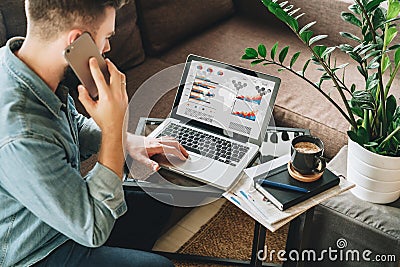 Young man, dressed in shirt, sits at home on couch at coffee table,uses laptop with graphs, charts, diagrams on screen Stock Photo