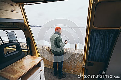 Young man drinking tea from thermos in front of camper van with a lake on background Stock Photo