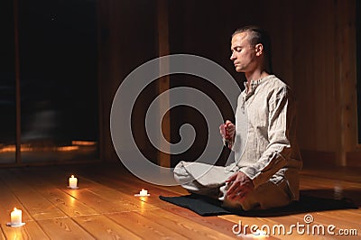 A young man in gray practice clothes sits in a lotus position with a red rosary in his hand in meditation. Dark room Stock Photo