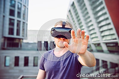 Young man with hand raised in front of him in virtual reality glasses headset standing against modern building background outdoors Stock Photo