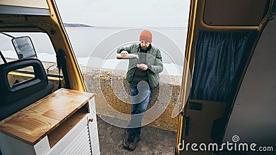 Young man drinking tea from thermos in front of camper van with a lake on background Stock Photo