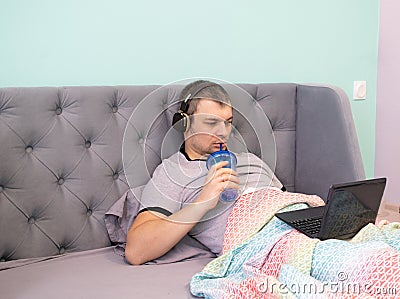 A young man in headphones lies reclining in a bed and watches video or works on a laptop. He holds a plastic glass with a tube and Stock Photo