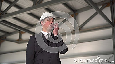 A young man in a helmet speaks on a walkie-talkie at a construction site. The boss in the suit looks around. Stock Photo