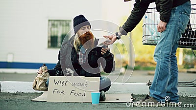 Young man helps to homeless person and giving him some food while beggar drink alcohol and sit near shopping cart at Stock Photo