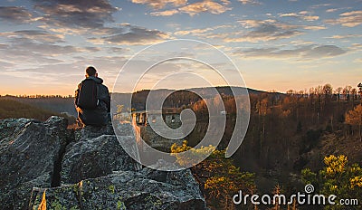 Young man in jacket, pants and backpack sitting and looking to ruin Divci kamen from rock. Czech republic Stock Photo