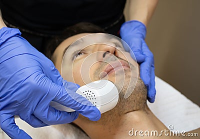 Young man laying on a facial therapy couch covered with the white towel with opened eyes. Facial therapist hands holding Stock Photo