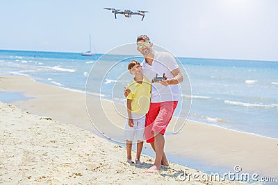 Young man with little boy flying drone at the sea shore, enjoying summer vacation. Stock Photo