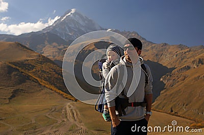 Young man with little child in baby carrier backpack near snowy mountain glacier at autumn, travelling with kids Stock Photo