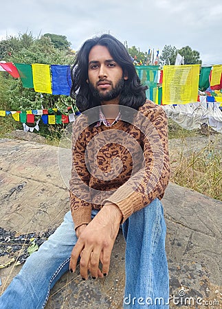 A young man with long hair and beard looking at camera while sitting over rock with the background of Tibetan prayer flags Stock Photo