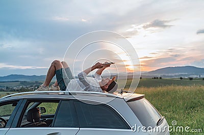 Young Man lying on the car roof and reading the paper bestseller book.He stopped his auto on the meadow with a beautiful valley Stock Photo