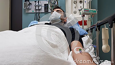 Young man lying in hospital bed. Recovering in modern hospital wards, covered with blanket, face mask and with intravenous needle Stock Photo