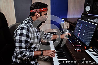 Young man, male artist looking focused while playing keyboard synthesizer, sitting in recording studio Stock Photo
