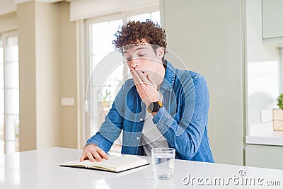 Young man reading a book at home cover mouth with hand shocked with shame for mistake, expression of fear, scared in silence, Stock Photo