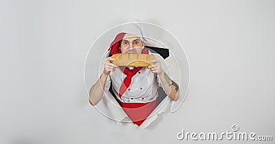 Young man with red dreadlocks dressed as chef bites white bread, standing in hole of white background. Hungry male cook Stock Photo