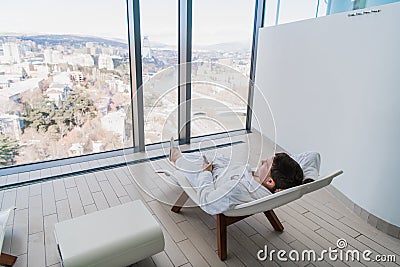 Young man resting on modern deck chair in front of big window with city view in modern spa Stock Photo