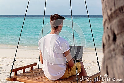 Young man seated on a swing and working with his laptop. Clear blue tropical water as background Stock Photo