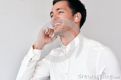 Young man smiling, having a call via wireless earphones with a colleague on white wall. Caucasian businessman wearing Stock Photo