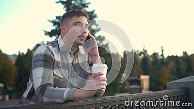 A young man speaks on the phone with a glass of coffee in his hand. A man with a phone near the fence on the embankment Stock Photo