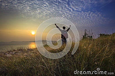 Young man standing and rising hand as victory on grass hill looking to sun above sea horizontal with dramatic colorful sky Stock Photo