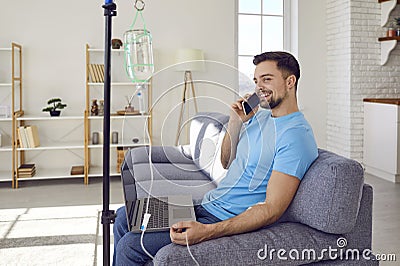 Young man talking by mobile phone sitting on sofa at home while receiving IV vitamin therapy. Stock Photo
