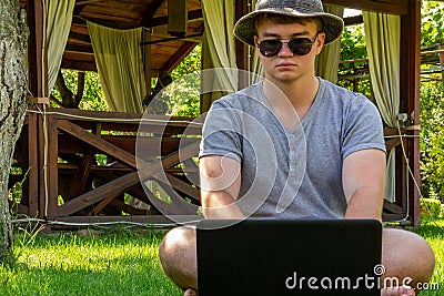 A young man, a teenager, a student, sits in a hat and sunglasses on the green grass, works on a laptop, against the background of Stock Photo