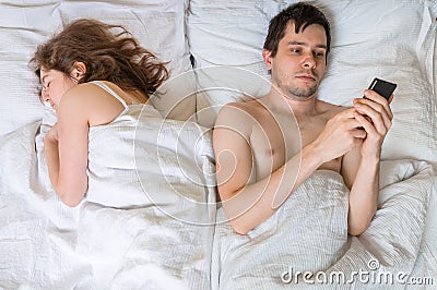 Young man is texting with someone using phone while his wife is sleeping near him. Stock Photo