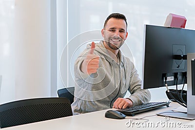 Young man with thumbs up happy and smiling for having achieved his goals in the office in front of the computer Stock Photo