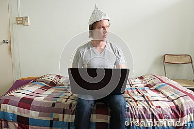 Young man with tin foil hat thinking while using laptop in the bedroom Stock Photo