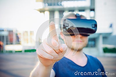Young man touching space in front of him in virtual reality glasses headset standing against modern building background outdoors. Stock Photo