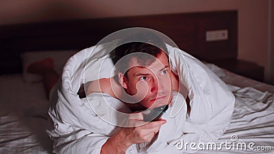 Young man under blanket lying on bed, watching tv and waving his legs in late evening. Relaxed male watching programs of Stock Photo